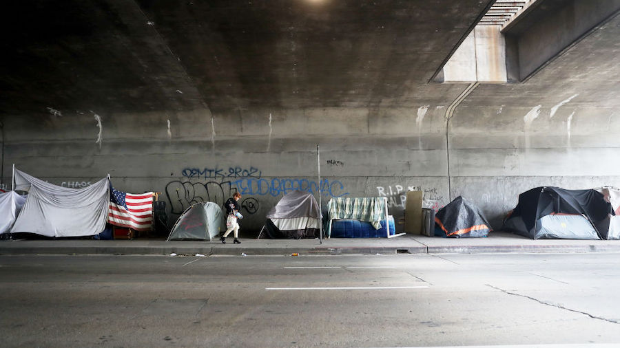 A woman walks past a homeless encampment beneath an overpass, with an American flag displayed, amid...
