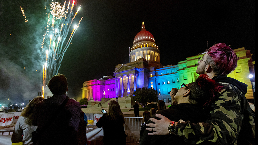 Fireworks shoot into the air next to the Idaho State House, which is illuminated in rainbow colors ...