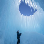 A person is looking up at a hole in the ice castle where the sky is visible. (AJ Mellor, Ice Castles)