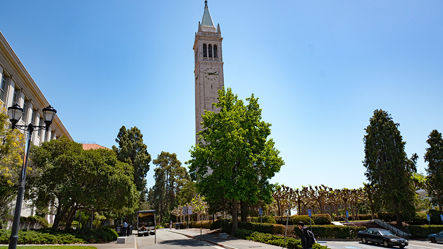 A young male student walks past Sather Tower (aka the Campanile) and other campus buildings on a su...