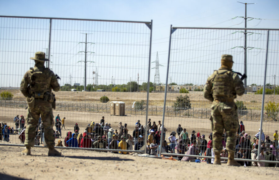 FILE - Migrants wait in line adjacent to the border fence under the watch of the Texas National Gua...