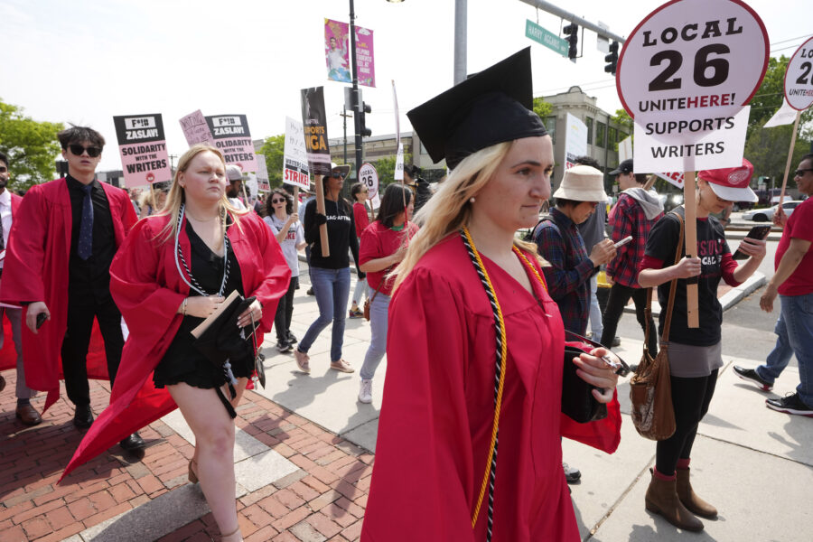 People dressed in commencement gowns, front, walk past protesters supporting the Hollywood writers'...