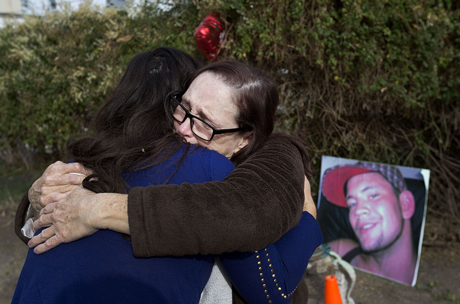 Two women hug in front of photo of man killed...