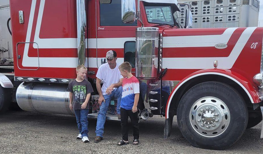 David Shultz in a baseball hat and jeans sits on a semi truck with two boys...