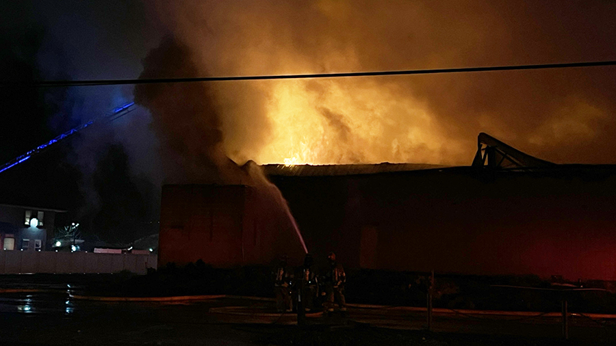 crews dump water on a roof as flames light up the smoke above it...